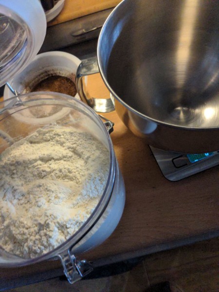 An open flour canister next to a mixing bowl on a scale. Behind them is the plastic dish I use to keep coffee grounds from getting everywhere when brushing off the coffee grinder, and which I probably should have moved before making a picture, but such is life.