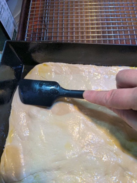 The dough, with a sheen of olive oil on it, now stretches to cover most of the bottom of the pan. A black silicone spatula is being used to press the dough towards the edges and corners of the pan, but the dough doesn't quite fill the corner.