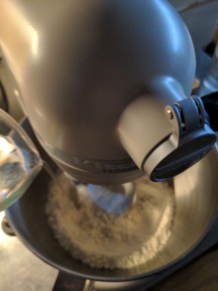 Water is slowly added to a mixture of flour, sugar, and yeast in the bowl of a KitchenAid stand mixer.