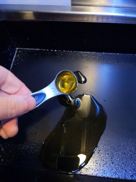 1½ tsp of olive oil is added to a pool of 1½ tsp of oil already in the bottom of a rectangular black metal pizza pan. The sides have been sprayed with cooking spray.